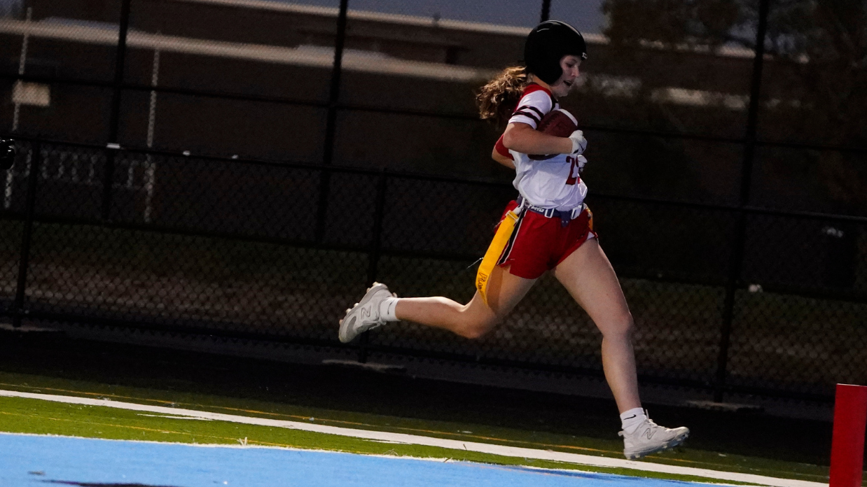 A person in a red and white uniform runs with a football towards the end zone on a field, wearing a helmet.