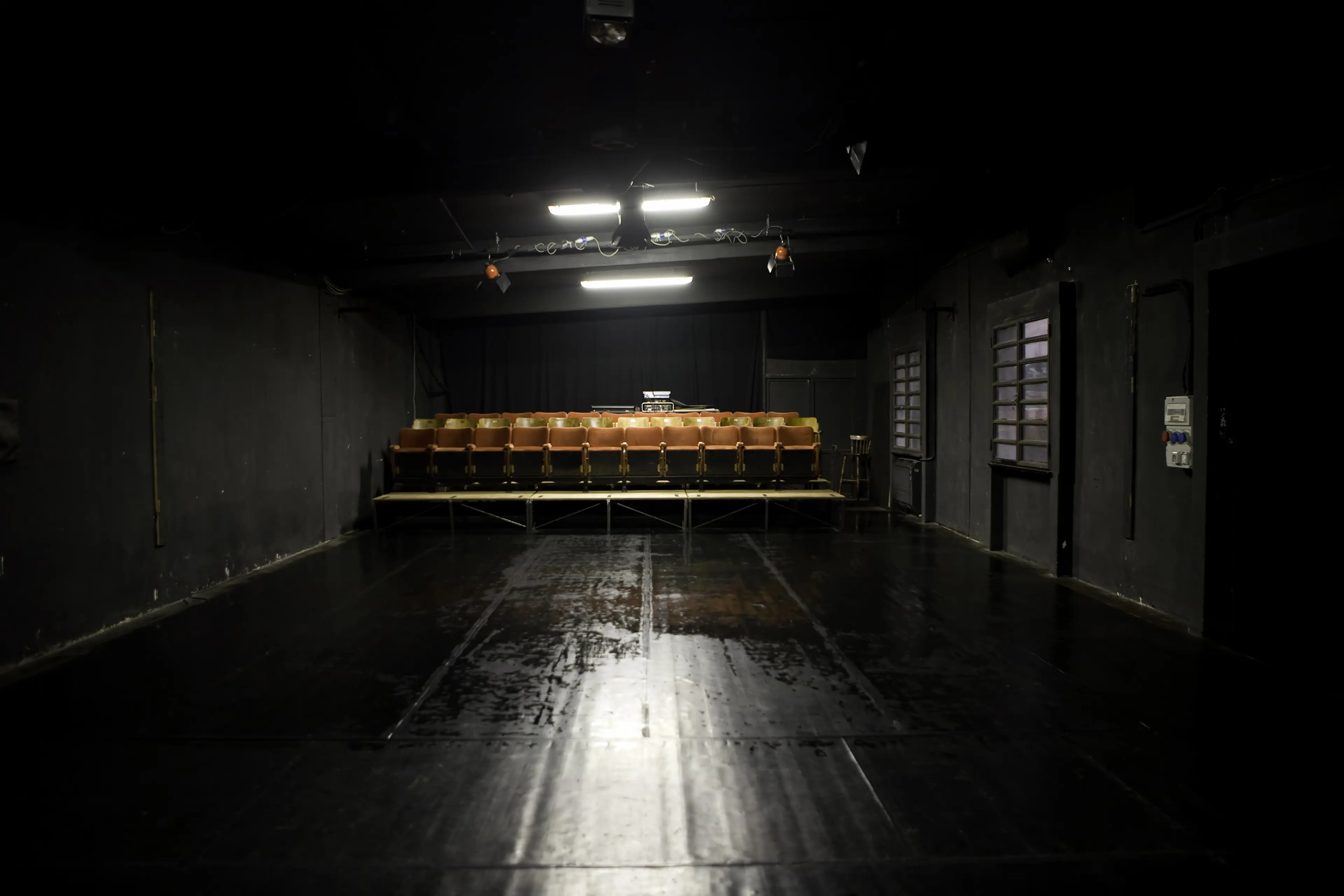 Dimly lit theater with a row of empty orange seats facing a dark, glossy stage floor. Overhead lights illuminate the seating area.