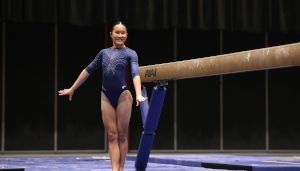 A gymnast in a blue leotard smiles confidently, standing beside a balance beam in an indoor arena.