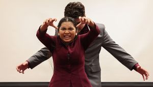A woman in a burgundy outfit stands in front of a man in a suit, making a fierce expression with her hands raised like claws.