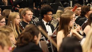 A diverse group of musicians in formal attire play woodwind instruments in an orchestra setting.