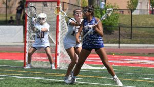 Two female lacrosse players in action near the goal, one in blue holding the ball, the other in white defending. A goalie stands in the background.
