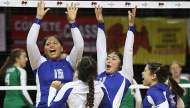 Four volleyball players in blue jerseys celebrate energetically with raised arms on the court. A net and opposing team in green are visible.
