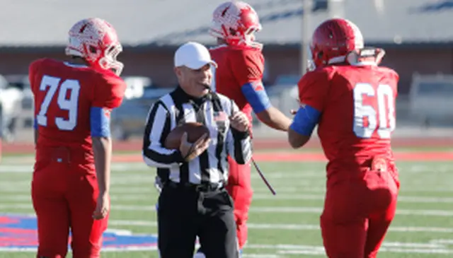 Referee holding a football talks to three American football players in red uniforms on a field. Players are wearing helmets with a feather design.