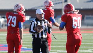 Referee holding a football talks to three American football players in red uniforms on a field. Players are wearing helmets with a feather design.