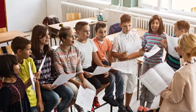 A group of children in a classroom sing while holding sheets of paper, led by a teacher with a music book.
