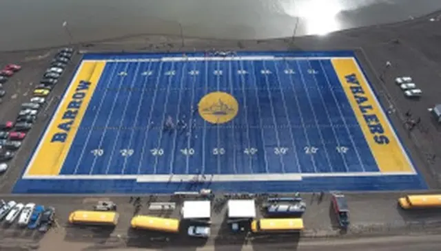 Aerial view of a blue and yellow football field labeled "Barrow Whalers," surrounded by parked vehicles and a body of water.