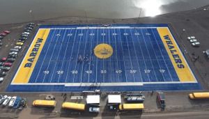Aerial view of a blue and yellow football field labeled "Barrow Whalers," surrounded by parked vehicles and a body of water.