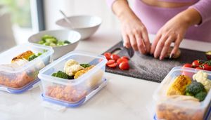 Person preparing meals with vegetables and grains, placing ingredients in containers on a kitchen counter, with salad in the background.