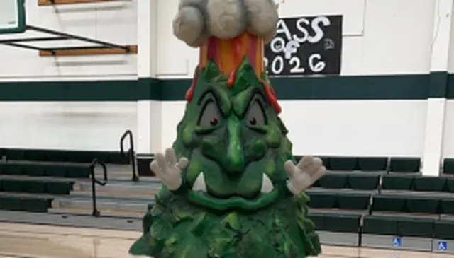A large, green tree mascot with an erupting volcano on top stands in a gymnasium, with "Class of 2026" sign in the background.