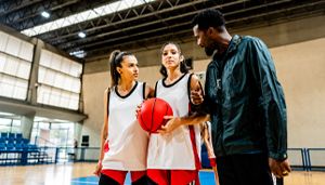 Two female basketball players holding a ball talk with an official on an indoor court, surrounded by natural light from large windows.