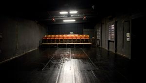 Dimly lit empty theater with black walls and floor, featuring a row of red seats on a raised platform at the back.