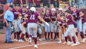 Softball team in maroon uniforms celebrates as a player wearing number 23 runs towards them. An umpire stands nearby on the dirt field.