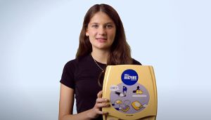 A person holding a yellow automated external defibrillator (AED) against a light blue background.