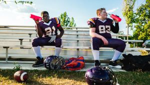 Two football players in purple uniforms sit on bleachers, holding red water bottles. Helmets and gear are on the grass in front of them.
