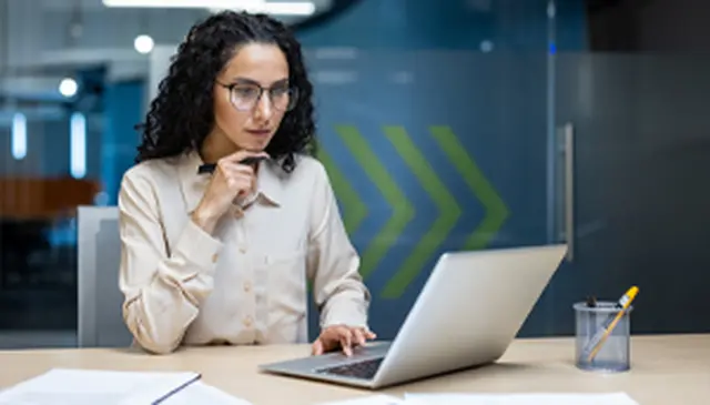 Woman with curly hair and glasses working on a laptop at a desk, holding a pen, in a modern office setting.