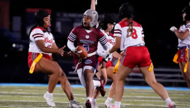 A flag football player in maroon tries to evade defenders in red and white uniforms on a field at night.