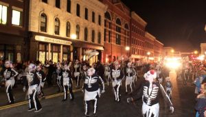 People in skeleton costumes parade on a city street at night, with historic buildings and lights in the background.