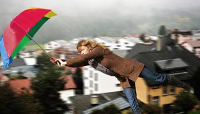 A person holding a colorful umbrella is lifted off the ground by strong wind, with a blurred cityscape in the background.