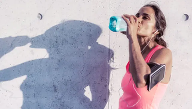 Woman in pink tank top drinking from a blue bottle, wearing earphones, with a smartphone armband, casting a shadow on a concrete wall.