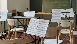 A music classroom with sheet music on stands, empty chairs, a keyboard, and a chalkboard in the background.