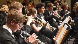 A group of musicians in formal attire plays brass instruments in an orchestra setting.