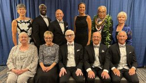 A group of people in formal attire pose together in front of a blue curtain, smiling. Some are seated, and others stand behind them.