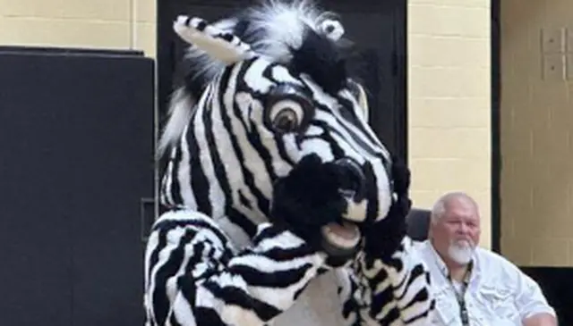 A person in a zebra costume stands indoors, holding their face with both hands. A seated man is visible in the background.