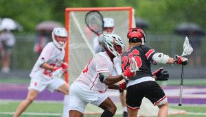 Lacrosse game in action; player in white defends against player in black near the goal, with teammates and goalie in the background.