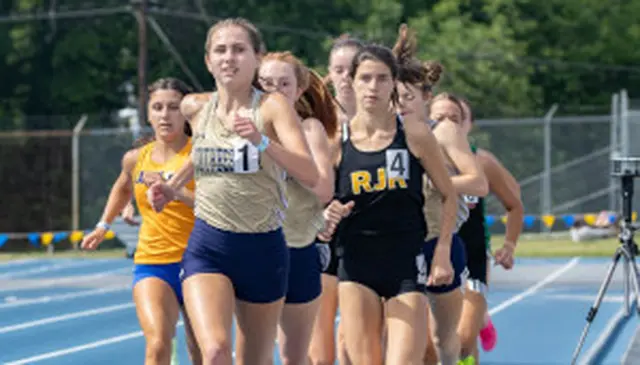 A group of female athletes running on a blue track during a race, with trees and a fence in the background.