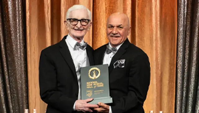 Two men in tuxedos smiling and holding an award plaque in front of a gold curtain backdrop.