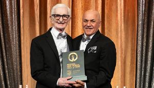 Two men in tuxedos smiling and holding an award plaque in front of a gold curtain backdrop.