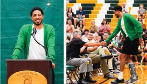 A man in a green cardigan speaks at a podium and shakes hands with a seated man in a gymnasium with a cheering crowd.