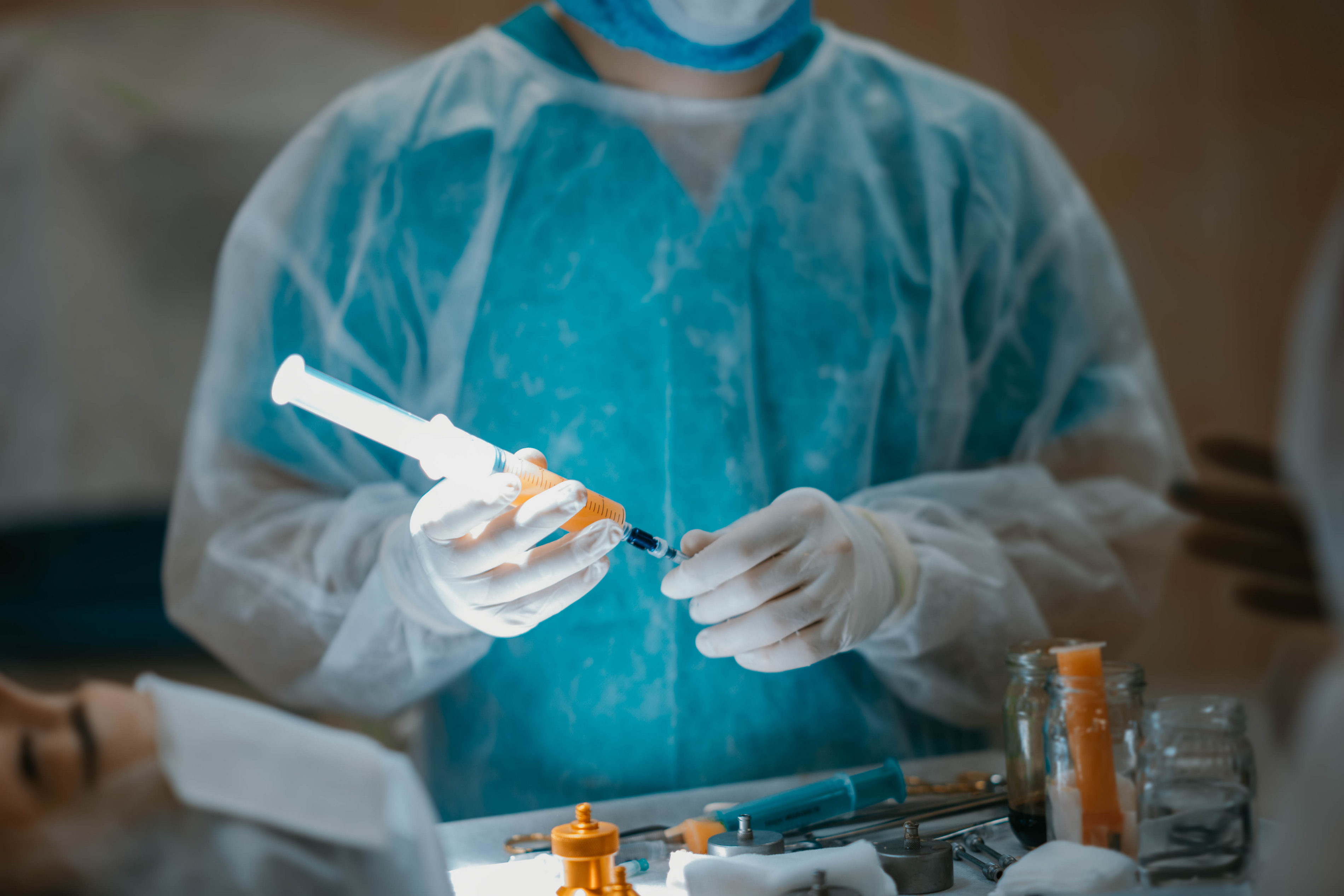 A medical professional in protective gear and gloves prepares a syringe amid surgical instruments, with a patient lying nearby.