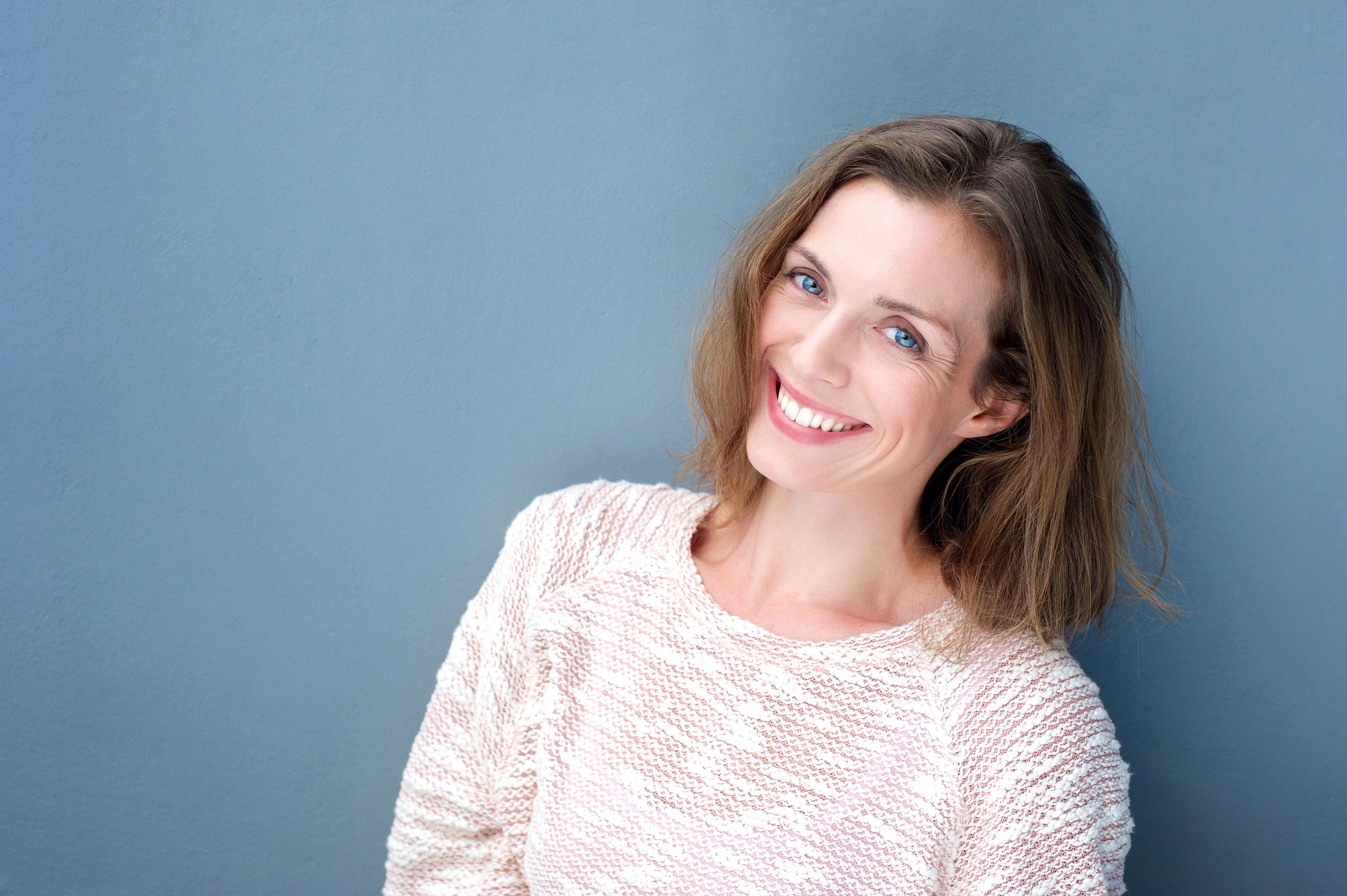 Woman with shoulder-length hair smiling, wearing a textured light sweater, against a plain blue background.