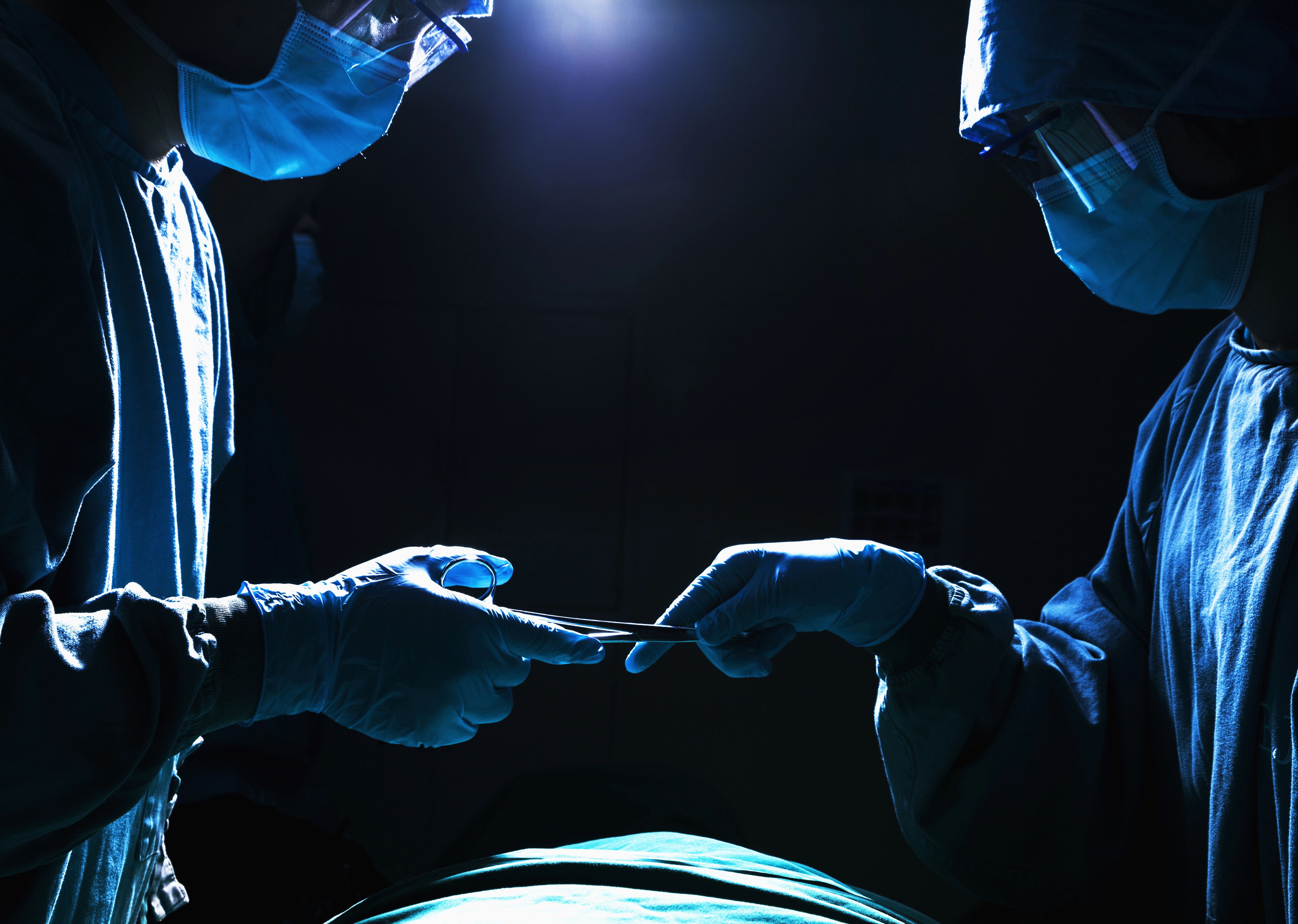 Two surgeons in dim lighting, wearing masks and gowns, exchanging a surgical instrument over a patient, illuminated by overhead light.
