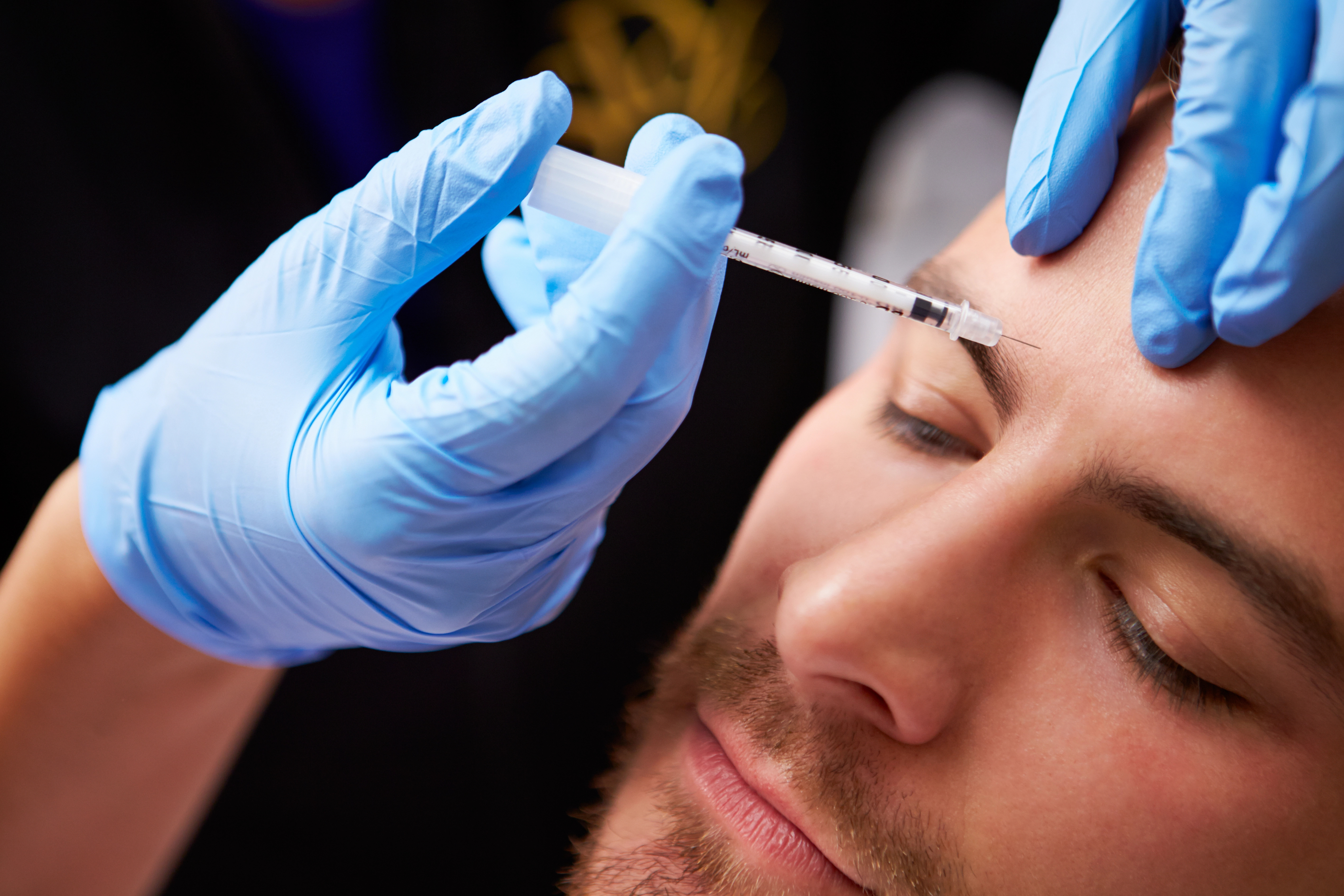 Person receiving a cosmetic injection in the forehead, administered by hands in blue gloves.