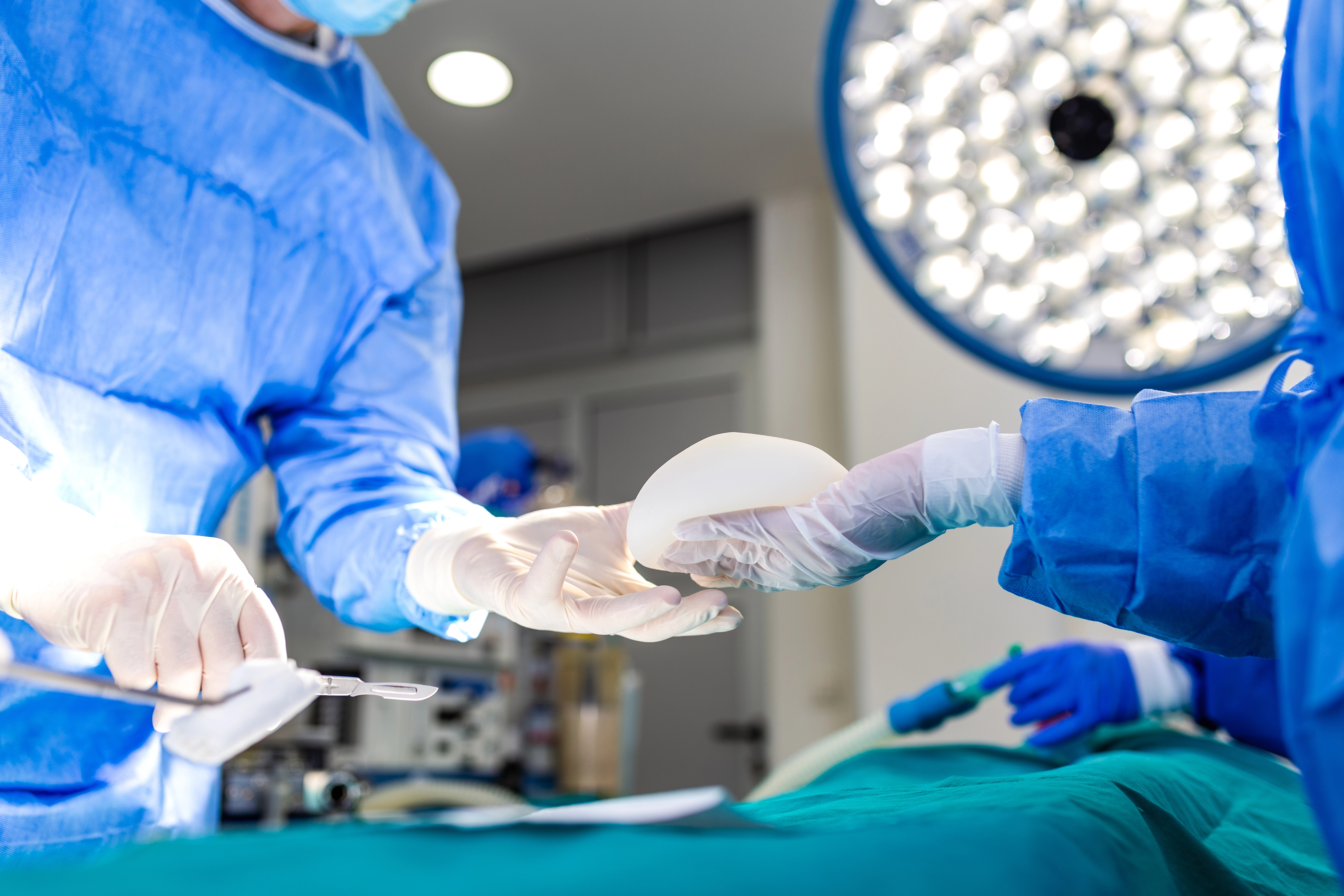 Surgeons in blue scrubs and gloves pass a breast implant in a brightly lit operating room.