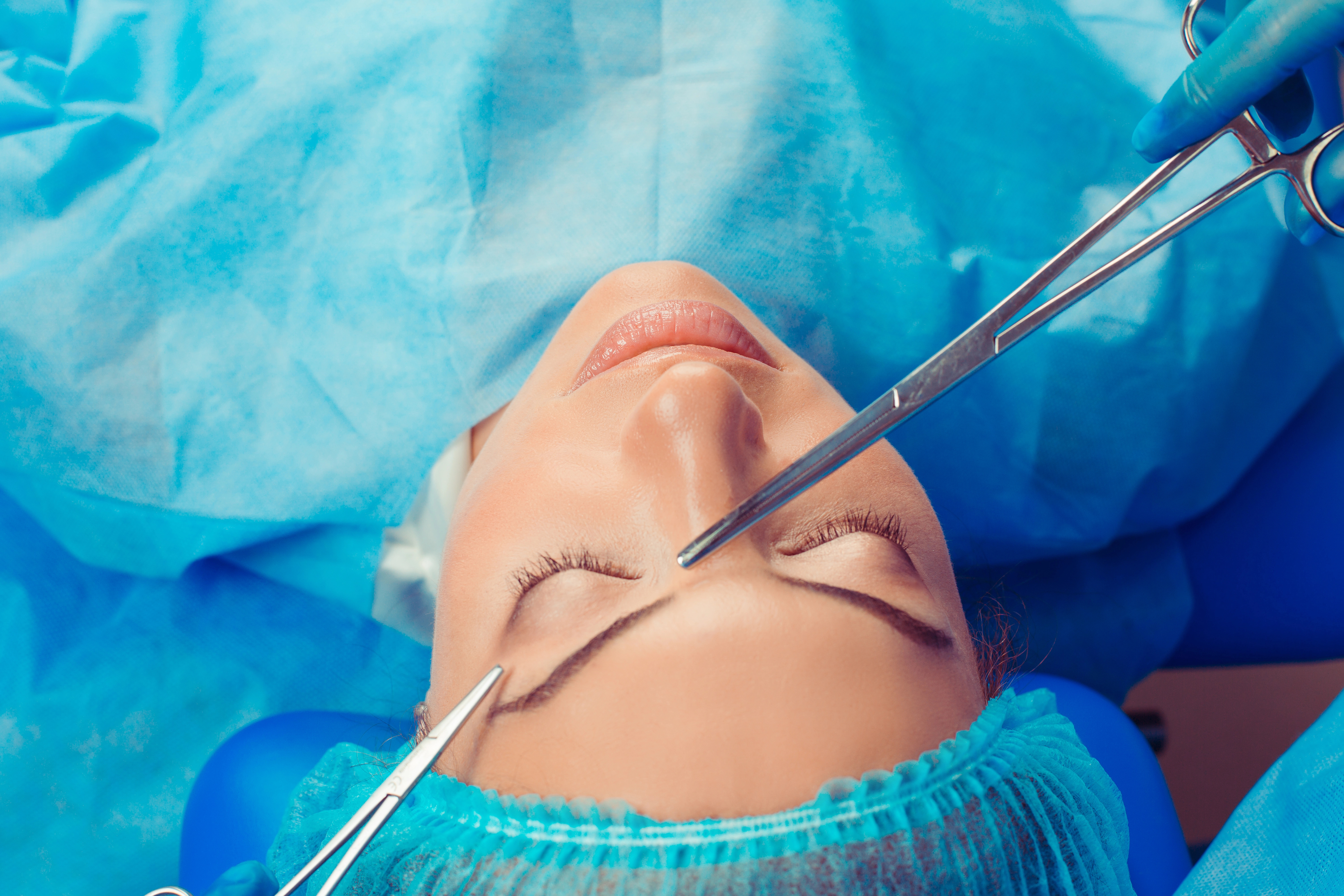 Person lying down with surgical tools near forehead, wearing a blue surgical cap and gown.