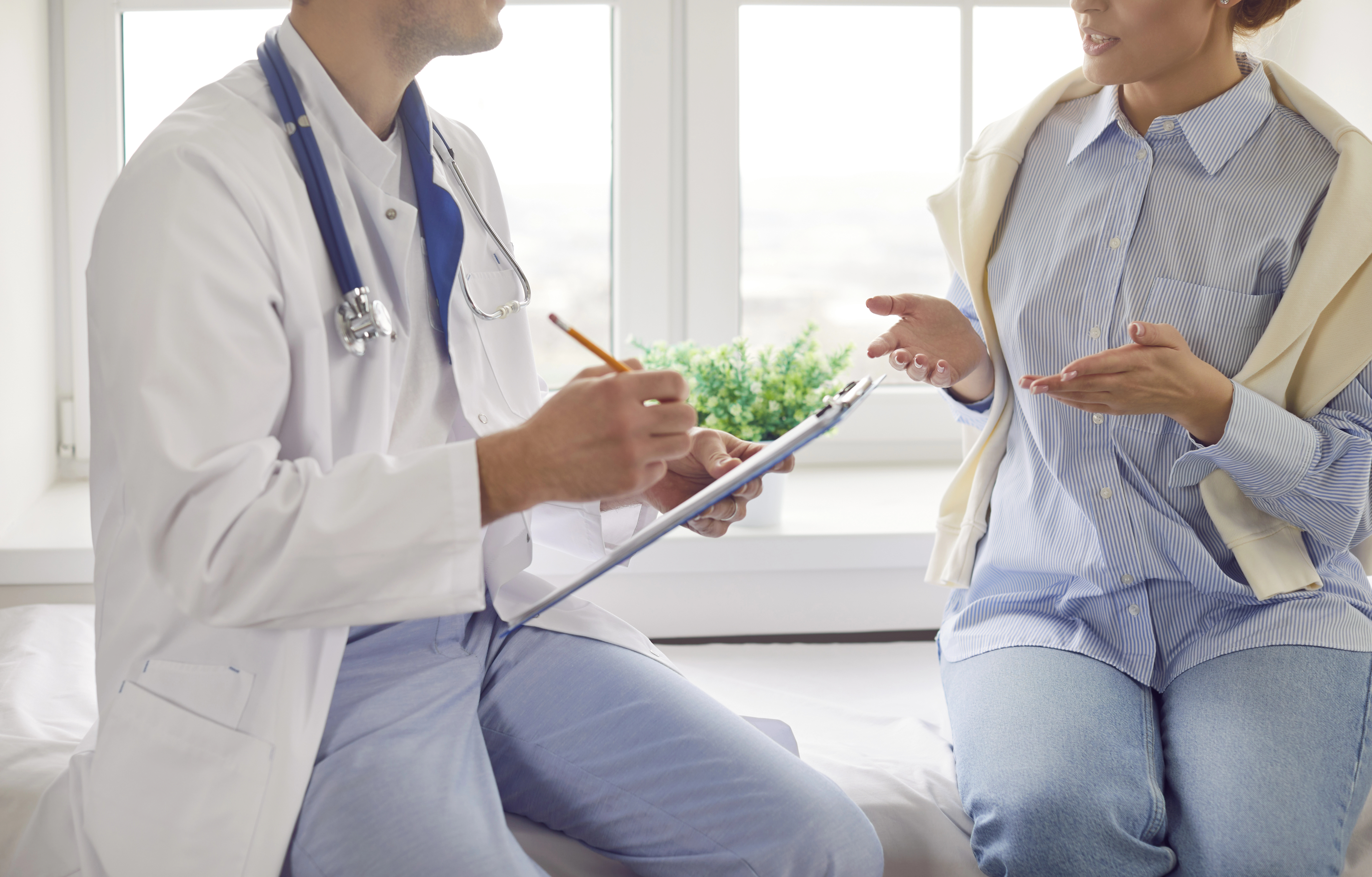 Doctor with stethoscope consults a patient in a light-filled room, taking notes on a clipboard as they discuss medical concerns.