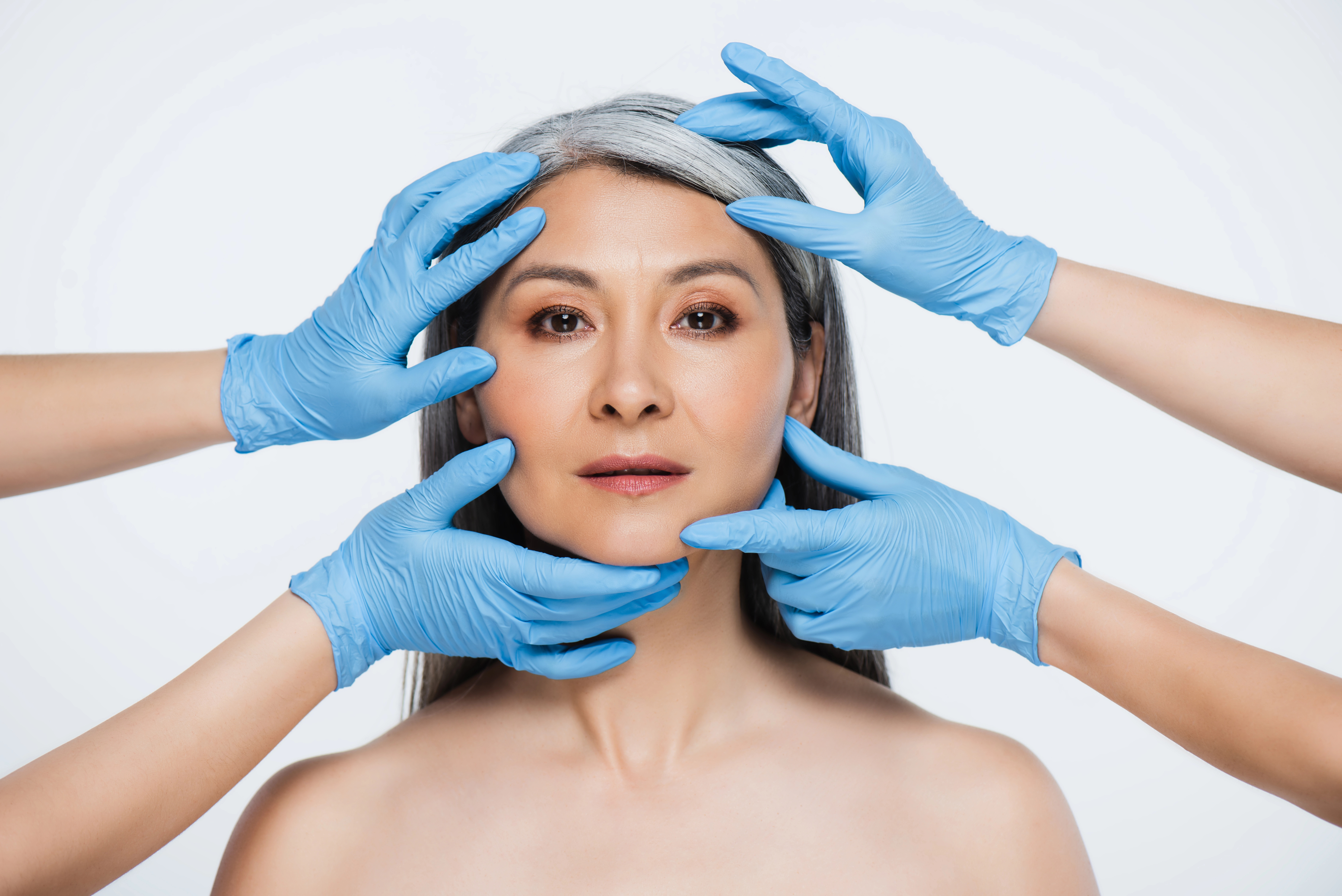 Woman with gray hair surrounded by blue-gloved hands adjusting her face, indicating cosmetic or dermatological examination.