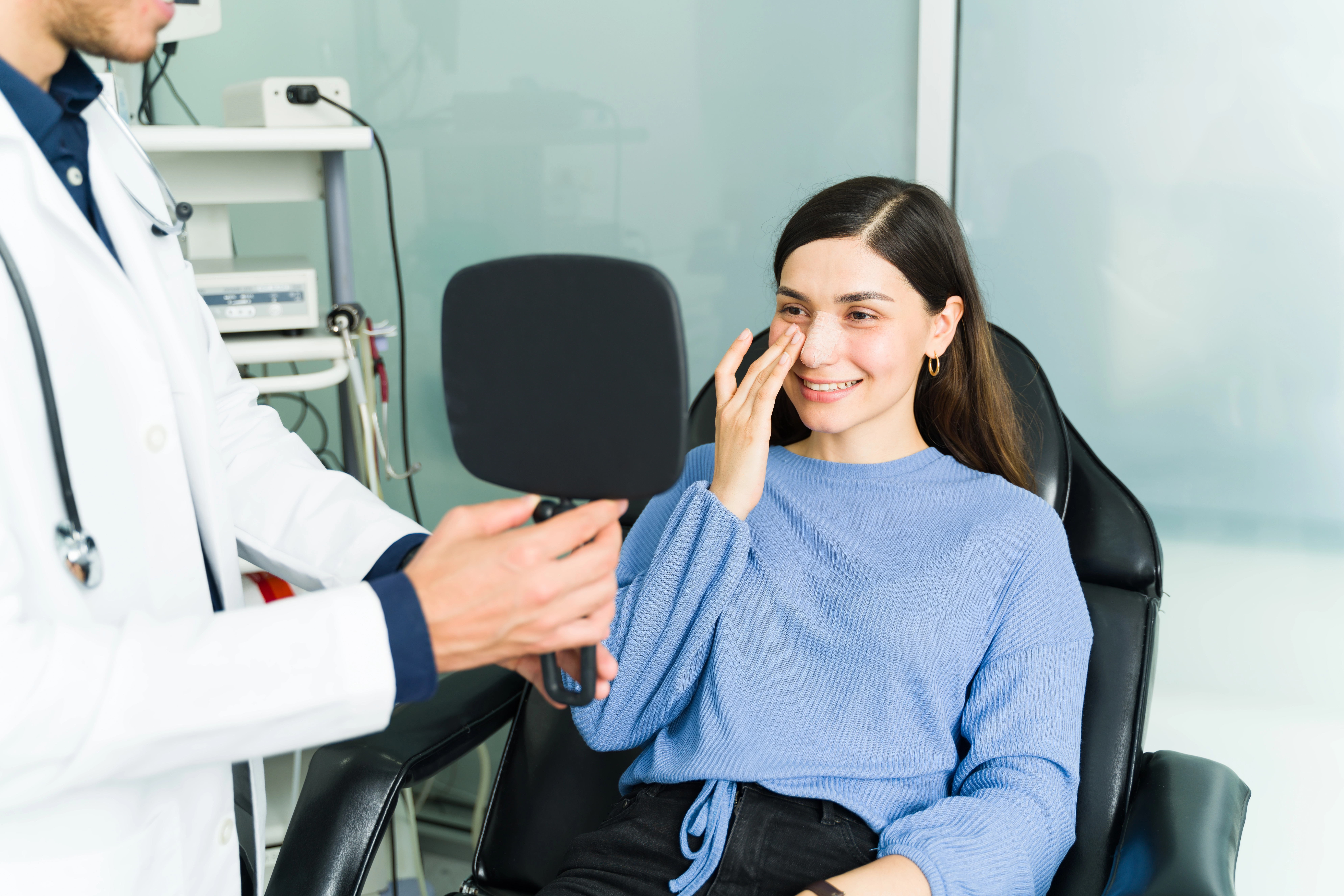 A woman in a blue sweater smiles, seated in a chair, looking at herself in a handheld mirror held by a doctor in a white coat.
