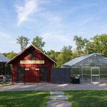Red barn labeled "Turtle Rock Farm" beside a greenhouse, surrounded by trees and a clear blue sky. Stone path leads to the buildings.