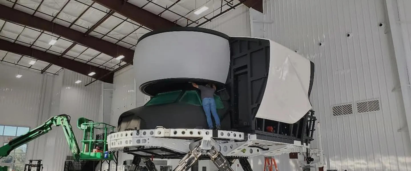 A person works on a large flight simulator inside a spacious, industrial facility with high ceilings and bright lighting.
