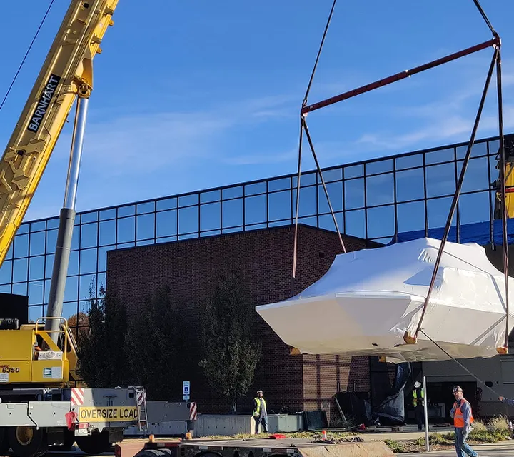 A crane lifts a large, wrapped flight simulator onto a flatbed truck outside a modern building, with workers guiding the process.
