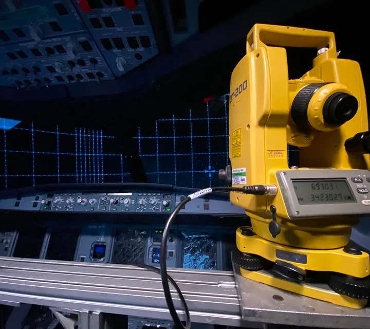 A yellow theodolite is set up inside a cockpit with illuminated control panels and a grid pattern displayed on a screen in the background.