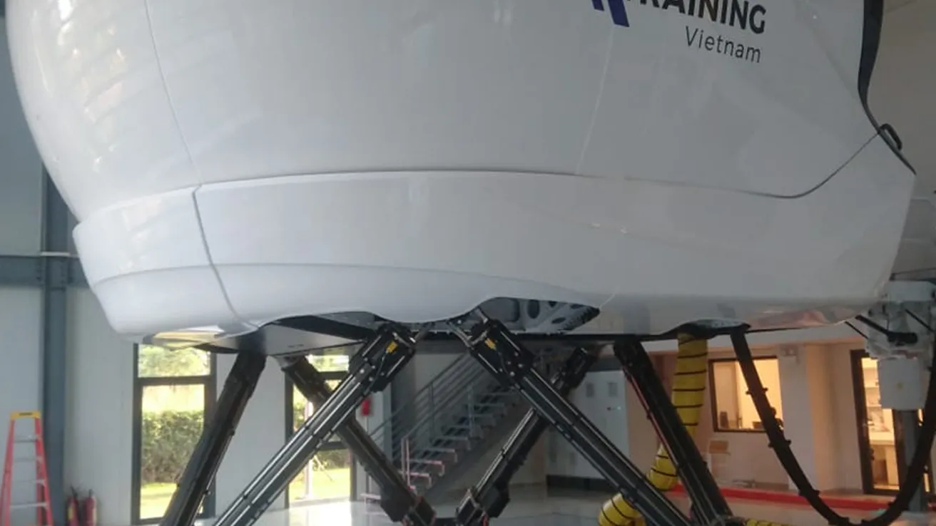 Flight simulator with "BA Training Vietnam" logo, mounted on hydraulic legs in a spacious hangar with glossy floors.