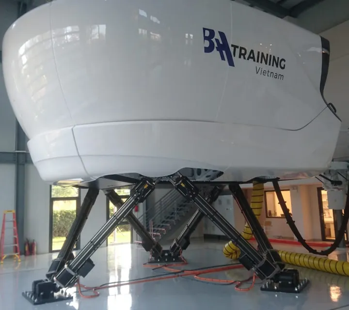 Flight simulator with "BA Training Vietnam" logo, mounted on hydraulic legs in a spacious hangar with glossy floors.