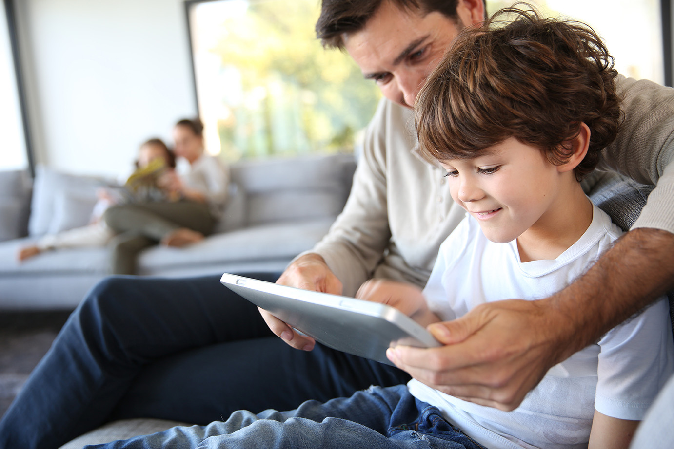 Photo of a father sitting on a sofa and guiding his young son as they look at a tablet computer together.