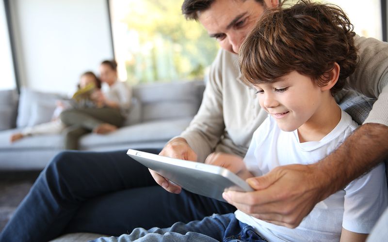 Photo of a father sitting on a sofa and guiding his young son as they look at a tablet computer together.